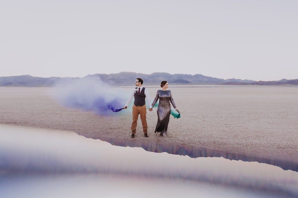 bride and groom pose on dry lake bed holding colorful smoke canisters