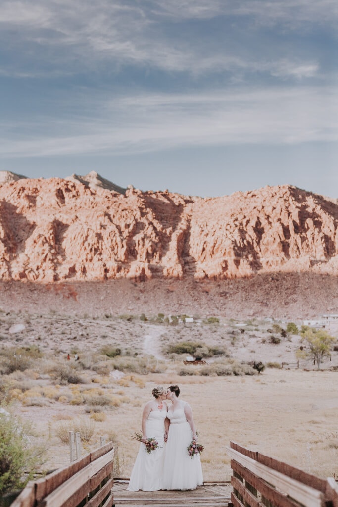 Two brides stand side-by-side kissing on the boardwalk of Calico Basin with Red Rock in the background.
