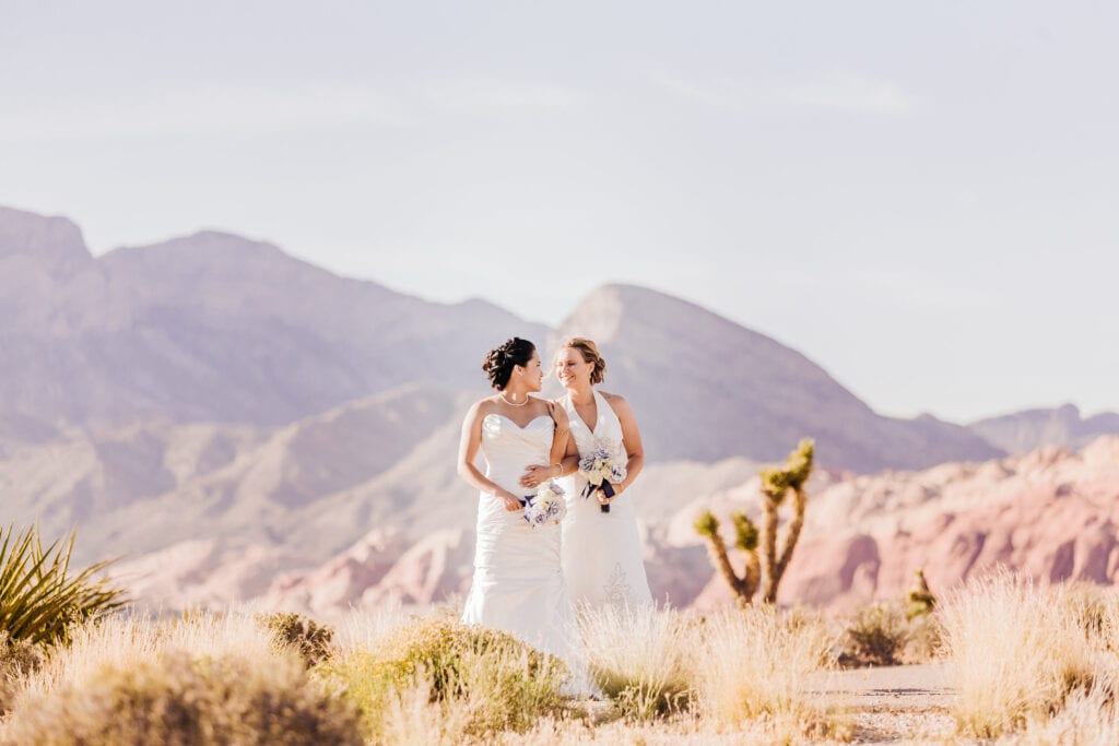 Two newlywed brides stand together laughing amongst Joshua Trees as Red Rock Canyon is seen in the background.