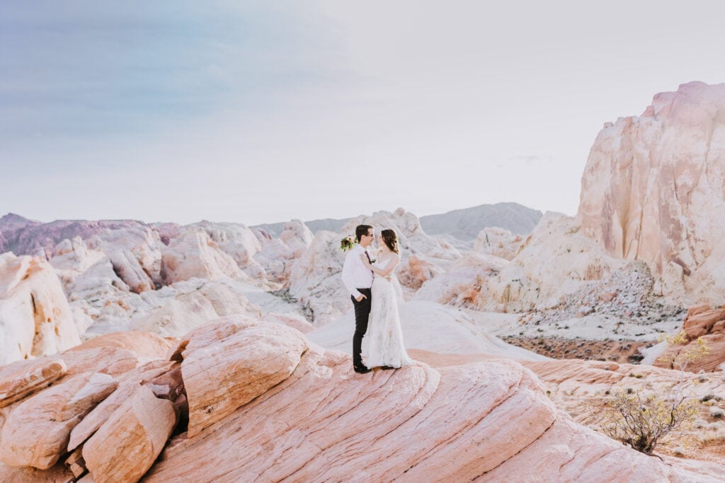 A wide shot of a couple standing atop a red rock with pastel cliffs in the background at Valley of Fire near Las Vegas.