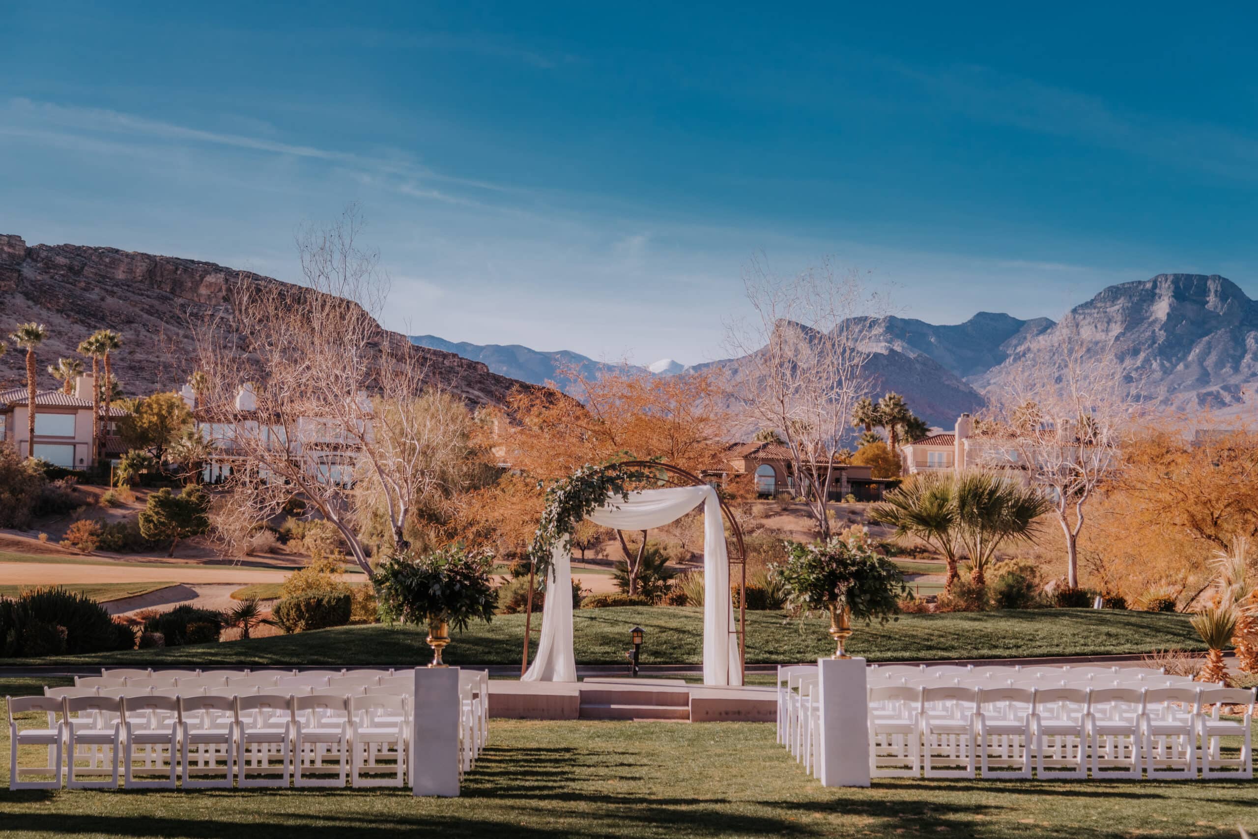 A wide angle view of a wedding ceremony setup with white chairs and white arch, with Red Rock as a backdrop at Red Rock Country Club in Las Vegas.