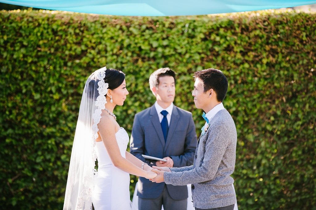 A bride and groom stand at the alter holding hands and saying their vows in front of a minister. They're all standing in front of a large green hedge.
