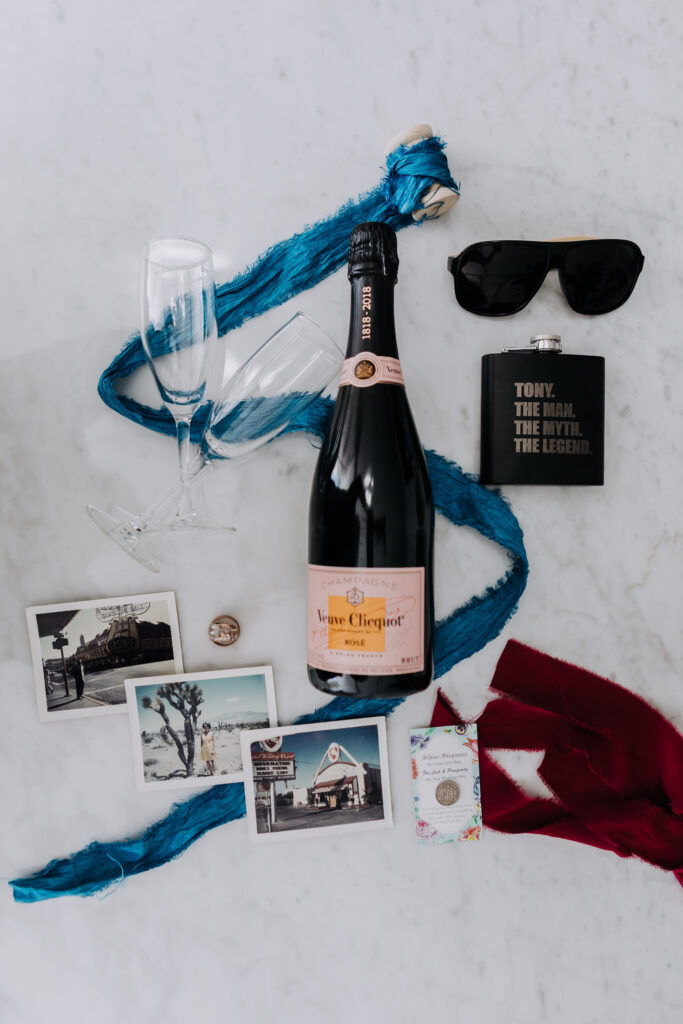 flat-lay of a champagne bottle with glasses, a flask, old polaroids of Las Vegas, and blue silk.