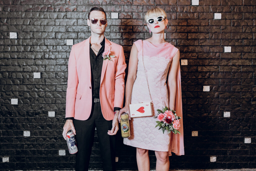 A couple pose against a black wall, dressed in black, pastel pink, and holding beers.