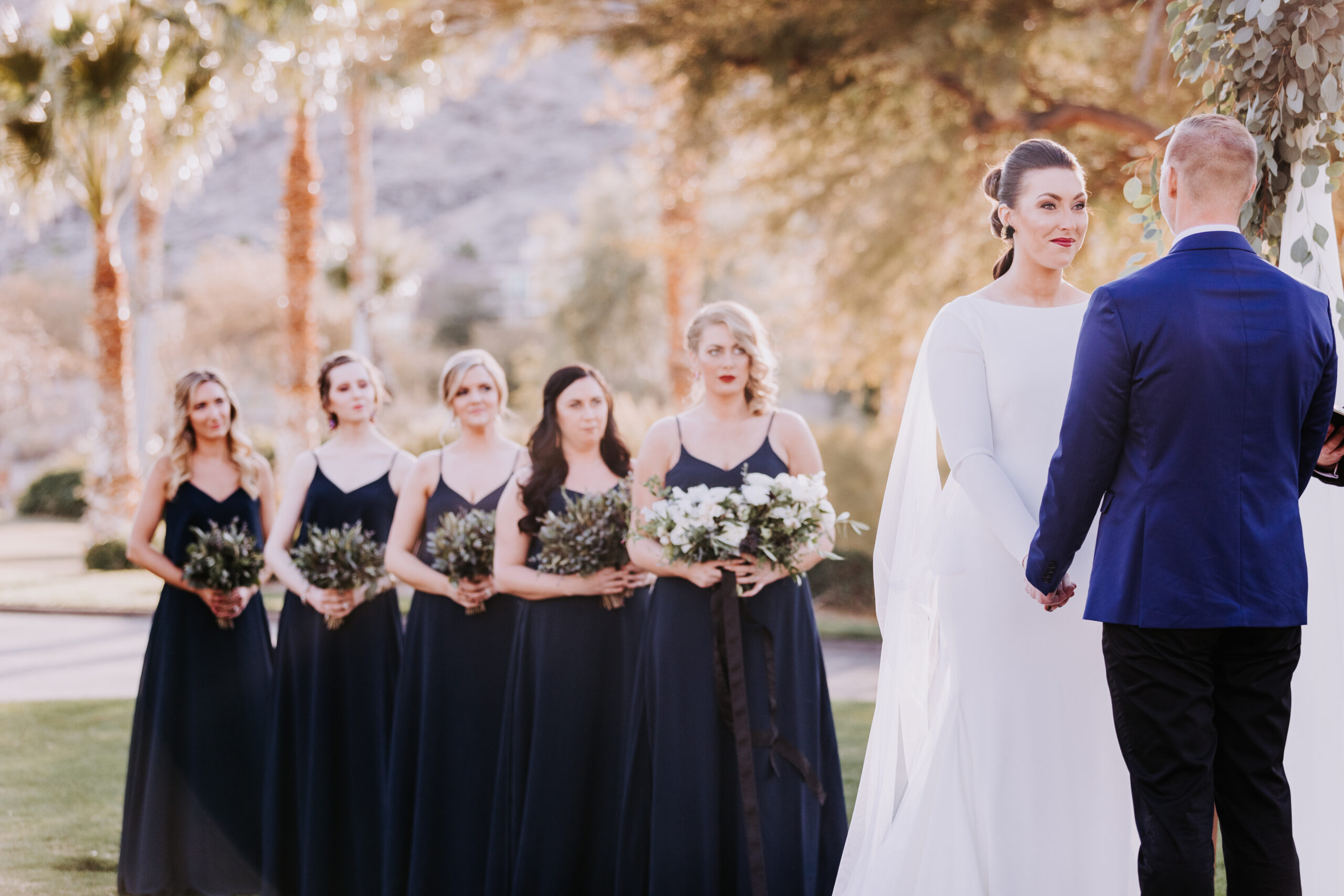 A bride listens to her groom's vows as several bridesmaids dressed in navy look on.