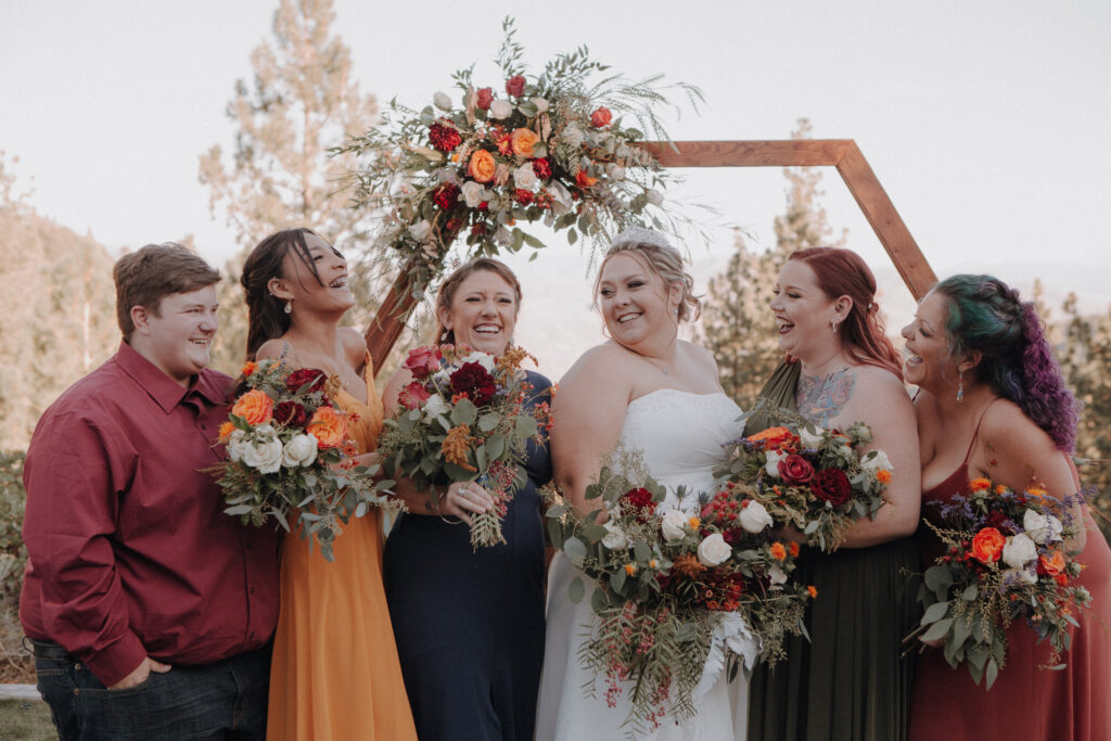A bride is surrounded by her bridesmaids who are all giggling together and dressed in vibrant blues, oranges, and burgandies.