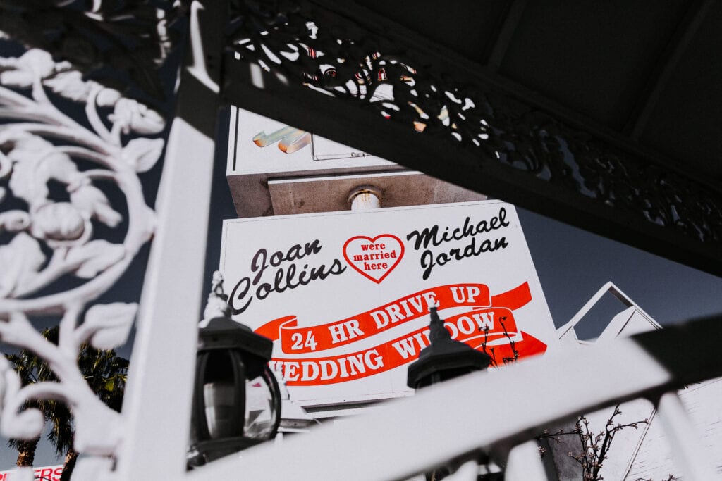 Looking upward, through a white gate, the bottom portion of a sign for A Little White Wedding Chapel is seen saying "Joan Collins and Michael Jordan were married here"