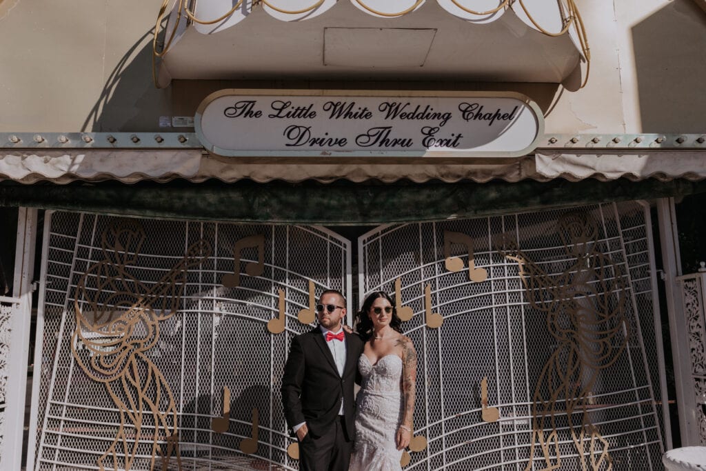A bride and groom pose in front of a white gate, adorned with golden musical notes, and wearing sunglasses under a sign denoting the entrance to a drive-thru at A Little White Wedding Chapel in Las Vegas.