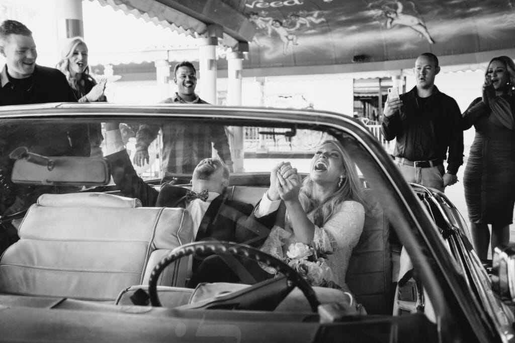 A black and white photo of a groom celebrating from the backseat of a vintage Cadillac after he says "I do" to his bride.