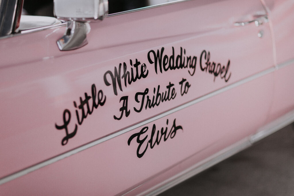 A close up image of the door of a Pink Cadillac that has "A Little White Wedding Chapel, a Tribute to Elvis" painted on it.