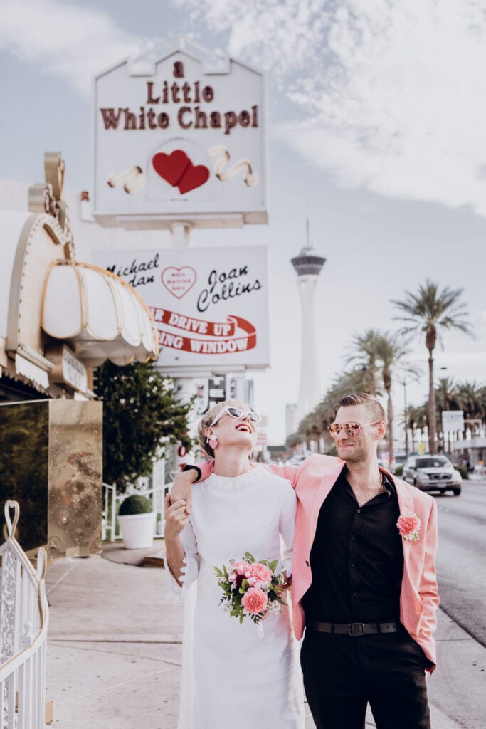 A bride laughs as her and her new husband walk down the street with a sign for A Little White Wedding Chapel in the background in Las Vegas.