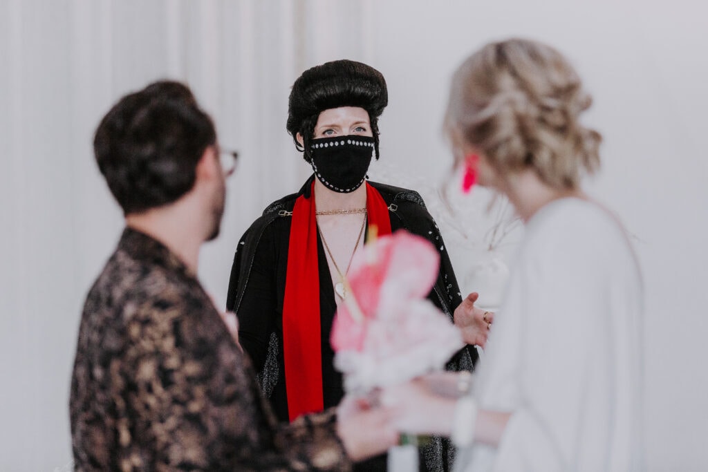 A female Elvis impersonator is in focus in the background, with a bride and groom in the foreground, as the Elvis impersonator officiates their wedding at Sure Thing Chapel in Las Vegas.